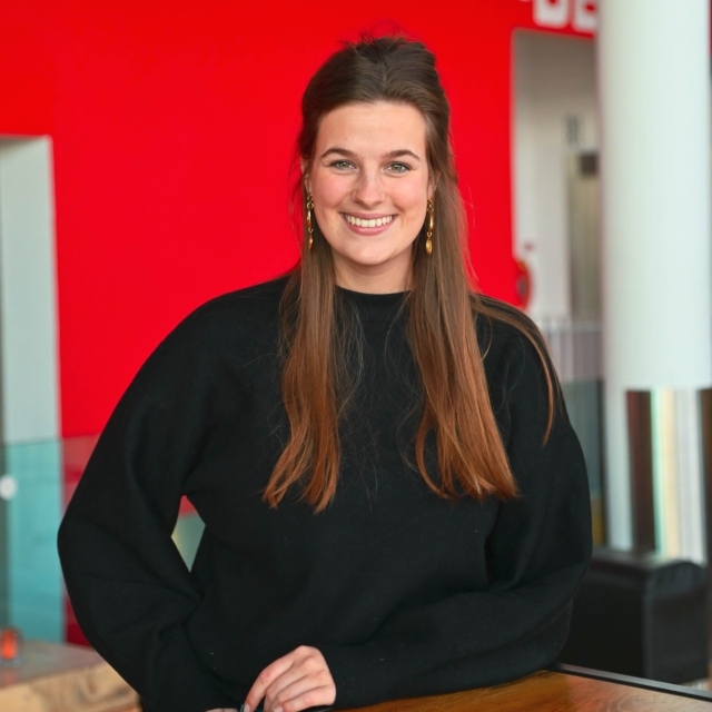 Vrouw met bruin lang haar en een zwarte trui kijkt lachend de camera in.