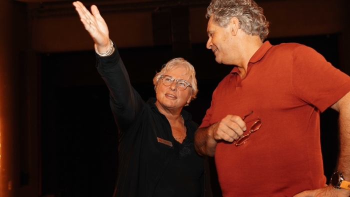 Een vrouw met kort licht haar en een bril heft haar arm omhoog en wijst terwijl ze naar een man met krullend grijs haar in een rood poloshirt kijkt, en de man houdt een bril in zijn hand en kijkt in dezelfde richting.