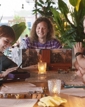 Een groep jonge mensen zit lachend rond een tafel in een caféachtige omgeving terwijl ze samen een bordspel spelen, met dobbelstenen, papieren en een verlichte kaars op tafel.