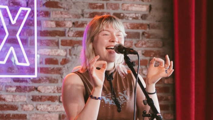 Een vrouw staat zingend achter een microfoon, met beide handen opgeheven in een gebaar, gekleed in een mouwloos topje en wijde broek, met een saxofoon naast haar.