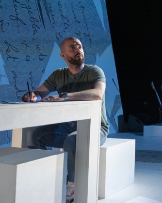 Derek Otte zit aan een witte tafel op een podium terwijl hij schrijft, met achter hem twee personen bij een laptop en muziekinstrumenten; de achtergrond toont een projectie van handgeschreven tekst in blauwe tinten.