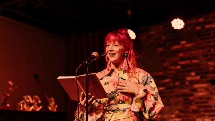 Een persoon met felroze haar staat voor een microfoon en leest uit een open notitieboek, gekleed in een fleurige blouse met grote bloemenprints, terwijl ze spreekt of voordraagt in Café Dox.
