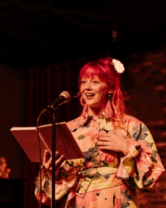 Een persoon met felroze haar en een kleurrijke bloemenjurk staat glimlachend op een podium en leest voor uit een notitieboekje bij een microfoon.