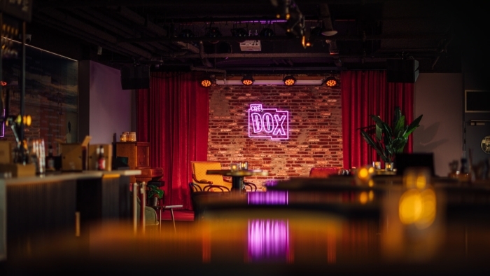 Een sfeervol café-interieur met centraal een bakstenen podiumwand waarop een felroze neonbord met de tekst “Café DOX” hangt, omringd door rode gordijnen, met tafeltjes, stoelen en warm gedimde verlichting op de voorgrond.