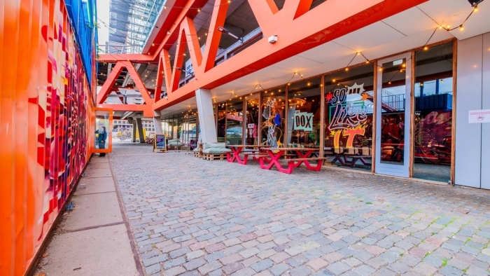 Een beschutte buitenruimte onder een fel oranje stalen constructie, met links een wand van kleurrijke graffiti en rechts glazen gevels met illustraties, waar houten picknickbanken en sfeerverlichting langs het geplaveide pad staan.