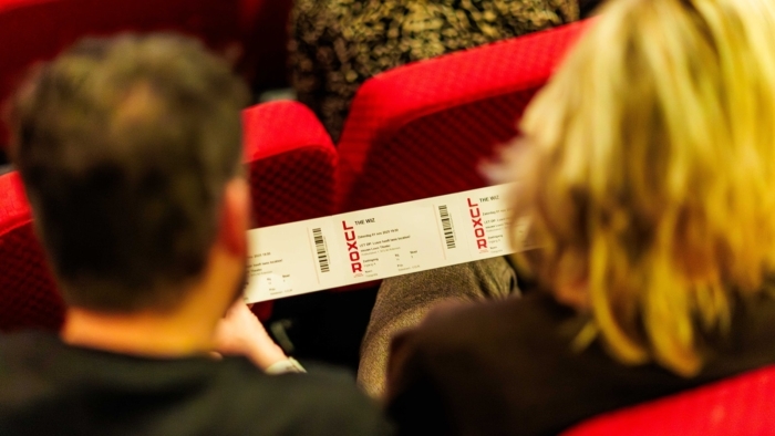 Twee personen die naast elkaar zitten in rode theaterstoelen, waarbij één van hen een set Luxor-toegangskaartjes met barcodes horizontaal voor zich houdt.