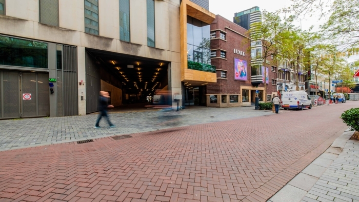 Een straatbeeld met links een groot, lichtgrijs gebouw met een brede verlichte doorgang, waar enkele personen lopend in beweging vervagen, en rechts het bakstenen gebouw van het oude Luxor met affiches aan de gevel en een busje dat langs de stoep geparkeerd staat.