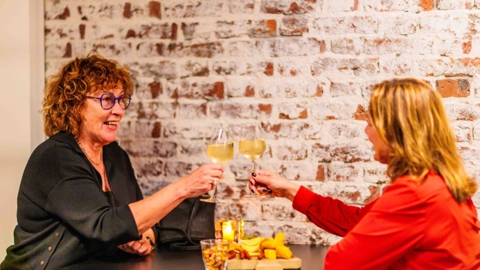 Twee vrouwen zitten tegenover elkaar aan een tafel terwijl zij elkaar aankijken en hun wijnglazen tegen elkaar tikken, met een borrelplank en een glas met nootjes voor hen op tafel.