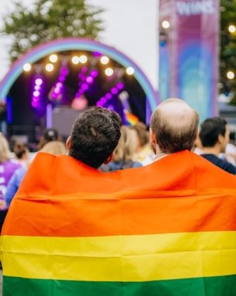 Twee personen staan met hun rug naar de camera en houden samen een regenboogvlag om hun schouders terwijl ze naar een podium kijken.